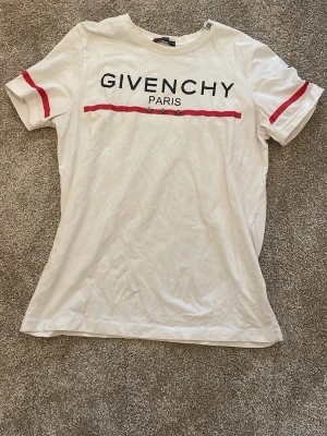 Vit Givenchy t-shirt med röd detalj - Säljer en vit t-shirt från Givenchy med svart logga och texten 'PARIS' framtill. T-shirten har röda ränder över bröstet och på ärmarna. Klassisk passform och rund halsringning. Perfekt för dig som gillar exklusiva streetwear-plagg.