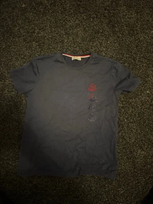 Mörkblå Moncler T-shirt - Snygg mörkblå t-shirt från Moncler med fyra loggor i rött och blått tryckta vertikalt på både fram- och baksidan. Klassisk rund halsringning och korta ärmar. Tillverkad i mjuk bomull för skön känsla och stilren look.