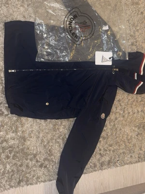 Marinblå vindjacka från Moncler - Snygg marinblå vindjacka från Moncler med dragkedja framtill och klassisk Moncler-logga på ärmen. Jackan har huva med rödvita ränder och diskreta sidofickor. Tillverkad i lätt, vattenavvisande material som passar perfekt till streetwear-stilen.  Storlek 3. Passar perfekt nu till våren. Vid funderingar skriv gärna ett meddelande mvh 47closet 😁