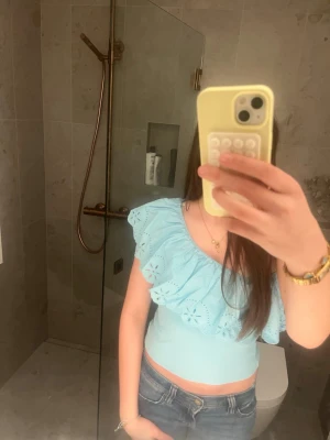 Ljusblå oneshoulder topp med volang - Säljer en ljusblå oneshoulder topp med stor broderad volangdetalj. Toppen är ribbad och har en snygg, tight passform som framhäver axlarna. Perfekt för dig som vill ha en unik och somrig look.