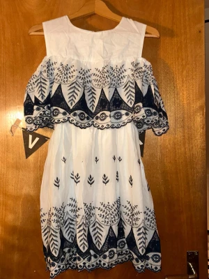 Vit off shoulder klänning med blå brodyr - Söt vit off shoulder klänning med blå broderade blad- och blommönster. Klänningen har volang upptill och vid kjol, samt fina detaljer längs kanterna. Perfekt för sommardagar och festivaler. Materialet känns lätt och luftigt.