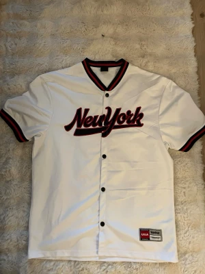 Vit baseballskjorta med röd text - Säljer en vit baseballskjorta med knappar och röd-svart text på bröstet. Skjortan har svarta och röda detaljer på krage och ärmslut, och är i en loose fit. Perfekt för dig som gillar sportig och avslappnad stil. Storlek S I herr modell 