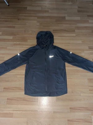 Nike Miller  - Snygg svart vindjacka från Nike med huva och dragkedja framtill. Jackan har två sidofickor med dragkedja och en vit Nike-logga på bröstet. Perfekt för blåsiga dagar och enkel att matcha med olika outfits.