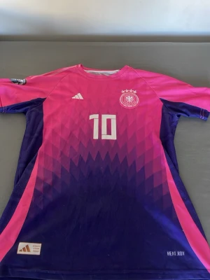 Rosa och lila fotbollströja Adidas - Snygg fotbollströja från Adidas i rosa och lila med nummer 10 på bröstet och Tysklands landslagslogga. Tröjan har coolt mönster, tre ränder på ärmen och HEAT.RDY-material som andas. Perfekt för träning eller match.