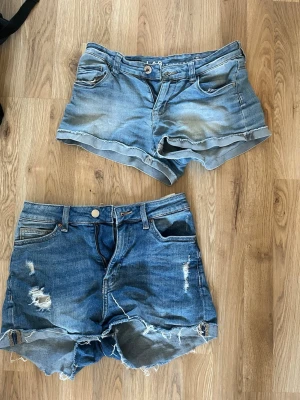 Två par blå jeansshorts - Två par blå jeansshorts, ett ljusare och ett mörkare par. Båda har klassisk femficksdesign, knapp och dragkedja fram. Det mörkare paret har slitna detaljer och fransig kant, medan det ljusare har uppvikta benslut. De mörkare är högmidjade och ger en fin passform medan det ljusare är lågmidjade. De mörka är st 36 och de ljusa st 158. Perfekta för sommaren!