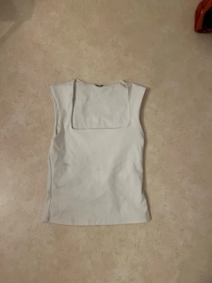 Vit ärmlös linnetopp - Snygg vit ärmlös linnetopp med fyrkantig halsringning. Enkel och stilren design som passar perfekt till jeans eller kjol. Toppen har en clean look och är lätt att matcha med andra plagg.