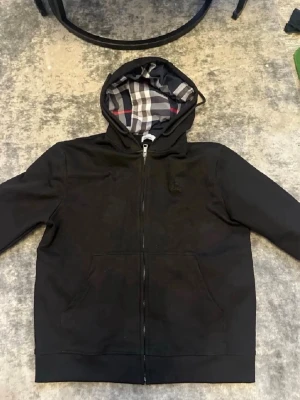 Svart hoodie från Burberry med dragkedja - Svart hoodie från Burberry med klassiskt rutigt foder i huvan och dragkedja framtill. Broderad Burberry-logga på bröstet och två fickor. Perfekt för dig som gillar stilrena och exklusiva plagg med streetkänsla.