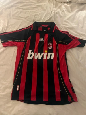 AC Milan matchtröja Kaka #22 Adidas - Säljer en klassisk AC Milan matchtröja från Adidas med Kaka och nummer 22 på ryggen. Tröjan är röd och svart med gula detaljer, klubbmärke och Bwin-tryck på bröstet. Perfekt för fotbollsfans som vill sticka ut på läktaren eller träningen. Tröjan är retro och därför extra speciell 