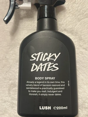 Lush Sticky Dates Body Spray 200ml - Sticky Dates Body Spray från Lush i en svart plastflaska med spraymunstycke. Doften är en blandning av benzoinresinoid och sandelträ, vilket ger en söt och varm känsla. Flaskan rymmer 200 ml och har en modern, stilren design.