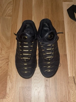 Nike Air Max Plus Tn Svart/Guld - •PRIS GÅR DISKUTERA•Nike Air Max Plus Tn sneakers i svart med guldiga detaljer och Tn-logga bak. Skorna har en sportig siluett, mesh- och syntetmaterial samt ikoniska vågformade linjer på ovandelen. Perfekta för dig som gillar streetwear och vill sticka ut med stilrena färger. 