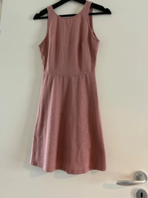 Rosa kortklänning med v-ringad rygg - Supersöt rosa kortklänning med rund halsringning fram och djup v-ringad rygg. Klänningen har en lätt utställd kjol och är ärmlös, vilket ger en fräsch och ungdomlig look. Perfekt för varma dagar eller sommarens alla tillfällen.