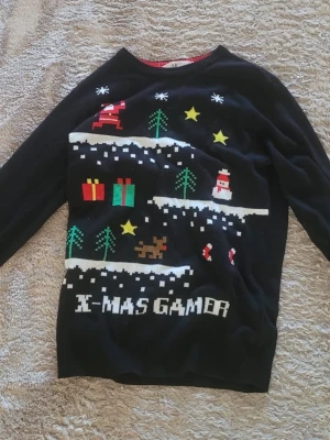 Svart jultröja X-MAS GAMER - Svart stickad jultröja med färgglatt pixelmönster av tomte, paket, stjärnor, snögubbe, granar och texten 'X-MAS GAMER' framtill. Perfekt för dig som älskar gaming och vill ha en unik jultröja. Rund hals och långa ärmar.