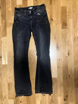 Svarta bootcut jeans - Snygga svarta bootcut jeans med slitna detaljer nertill och vita kontrastsömmar. Jeansen har klassisk femficksdesign och låg midja. Perfekta för en avslappnad och trendig look.
