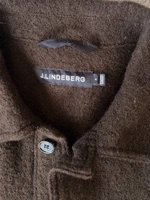 Brun overshirt från J.Lindeberg - Mörkbrun overshirt från J.Lindeberg i ullmix med klassisk krage, knäppning framtill och två stora fickor. Overshirten har en avslappnad passform och en mjuk, borstad yta som ger en cool och stilren look. Storlek S men känns som XS i längden!!