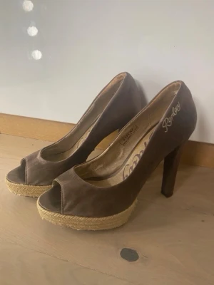 Bruna peep-toe pumps från Replay - Snygga bruna pumps från Replay med peep-toe och hög klack. Skorna har en platåsula i flätad jute som ger en cool espadrille-känsla. Ovandelen är i textil och klacken är smal för en elegant look. Perfekta för dig som vill sticka ut lite extra.