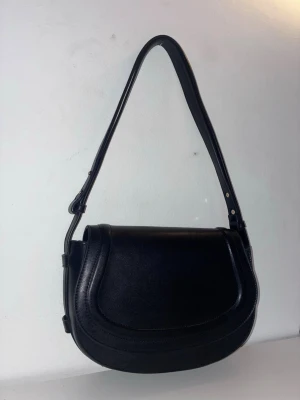 Svart axelväska i läderlook Kappahl - Stilren svart axelväska med rundad form och bred justerbar axelrem från Kappahl. Väskan har en slät yta i läderimitation och minimalistisk design utan synliga loggor. Perfekt för dig som gillar en clean och tidlös look. Aldrig använd.