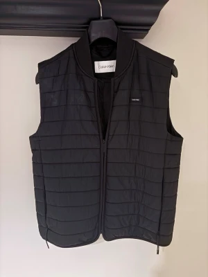 Svart dunväst från Calvin Klein - Stilren svart dunväst från Calvin Klein med quiltad design och diskret logga på bröstet. Västen har dragkedja framtill och en klassisk, rak passform. Perfekt att slänga över en hoodie eller tröja för extra värme och stil.