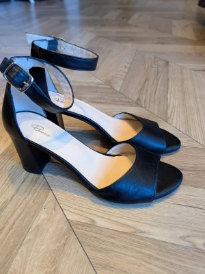 Svarta pumps med ankelrem och klack - Stilrena svarta läder pumps från Rizzo med öppen tå, bred klack och justerbar ankelrem med spänne. Skorna är tillverkade i slätt konstläder och har en bekväm innersula. Perfekta för dig som gillar klassisk och enkel design.