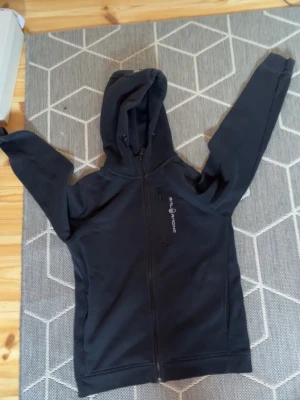 Svart hoodie från Sail racing - Snygg svart hoodie från Sea Ranch med dragkedja framtill och huva. Tröjan har en diskret logga på bröstet och en ficka med dragkedja. Perfekt för en avslappnad stil och enkel att matcha med andra plagg. pris går att diskutera. passar allt från 160-167