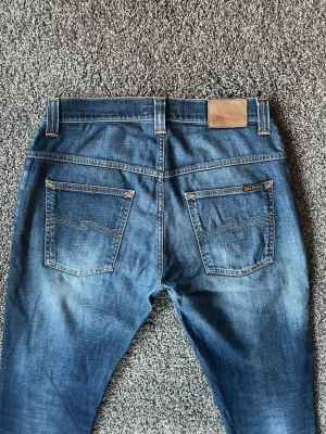 Blå nudie jeans - Säljer ett par blå straight fit jeans med klassisk femficksdesign och snygg läderpatch bak i midjan. Jeansen har kontrastsömmar och lätt slitning på benen för en cool look. Perfekta för dig som gillar en modern och avslappnad stil.