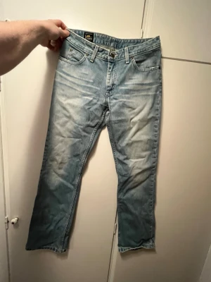 Ljusblå raka Lee jeans - Säljer ett par ljusblå jeans från Lee med klassisk femficksmodell och raka ben. Jeansen har slitna detaljer och en avslappnad passform, perfekta för en casual look. Materialet är mjuk denim i bomull och de har bälteshällor och knappgylf.
