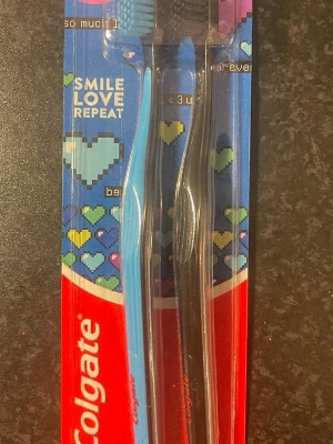 Colgate tandborstar 2-pack - Två-pack tandborstar från Colgate, en blå och en svart. Ergonomiskt formade handtag och mjuka borst. Perfekt för daglig tandvård. Förpackningen har färgglada hjärtan och texten 'Smile Love Repeat'.
