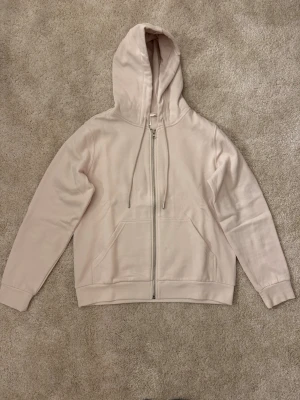 Ljusrosa zip hoodie med huva - Säljer en stilren ljusrosa hoodie med dragkedja framtill och stor ficka på magen. Hoodien har justerbar huva med snören och ribbade muddar vid ärmar och nederkant. Har en jätteliten fläck - se bild 4.