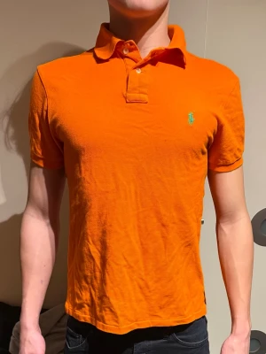 RALPH LAUREN PIKÉ - Säljer en orange pikétröja från Ralph Lauren med klassisk krage och knappar framtill. Tröjan har korta ärmar och den ikoniska gröna loggan broderad på bröstet. Perfekt till jeans eller shorts för en avslappnad men snygg look.                                          (Modellen är 180cm, 72kg)