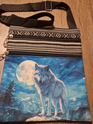 Axelväska med vargmotiv och mönster - Snygg axelväska i tyg med justerbar svart axelrem. Framsidan har ett coolt vargmotiv mot en blå natthimmel med fullmåne, och ovandelen har svartvita etnomönster. Bredd 19 cm. Höjd 23,5 cm. Väskan har två dragkedjefack och är perfekt för dig som gillar unika accessoarer.