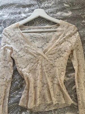 Krämvit spetsblus med v-ringning - Romantisk krämvit blus i spets med v-ringning och långärmad design. Blusen har fina volangdetaljer vid midjan och en feminin, figurnära passform. Perfekt för dig som gillar en söt och trendig stil.