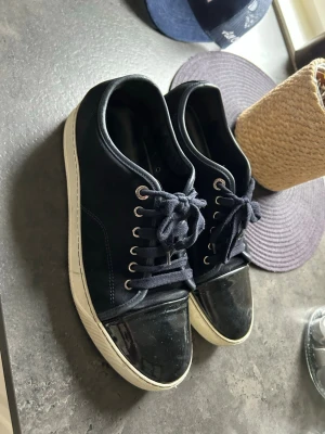 Lanvins! - Snygga svarta sneakers från lanvin  med coola lackdetaljer på tån och klassisk vit sula. Skorna har svarta skosnören, metallfärgade öljetter och en stilren design i både mocka och läder. Perfekta för dig som gillar en clean och modern look.