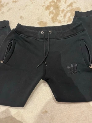 Svarta mjukisbyxor från Adidas - Svarta mjukisbyxor från Adidas med broderad logga på benet. Byxorna har dragsko i midjan, muddar vid bensluten och dragkedjor på fickorna. Sköna och stilrena, perfekta för chill eller träning. Har även en matchande tröja till ( kostar tillsammans 400kr )