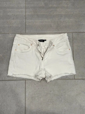Vita jeansshorts från Kappahl - Snygga vita jeansshorts från Kappahl med slitna kanter och klassiska fickor. Shortsen har hällor för skärp och stängs med dragkedja och knapp. Perfekta för sommaren och lätta att matcha med allt. Midwaist.