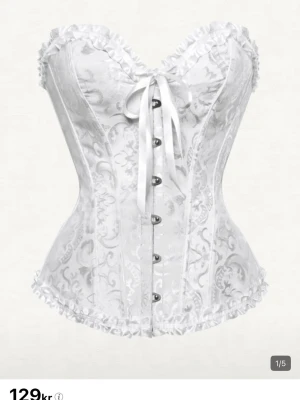 Vit spetskorsett med rosett - Elegant vit korsett med vackert mönstrad spets, dekorativa knappar framtill och en söt rosett vid bysten. Korsetten har volangkant upptill och nedtill samt markerad midja för en feminin siluett. Perfekt för dig som vill ha en romantisk och stilren look. Skriv om ni har några funderingar ❤️