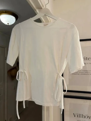 Vit topp med knyt i midjan - Vit topp med rund hals och korta ärmar. Toppen har snygga knytband i midjan på båda sidor som ger en trendig och justerbar passform. Perfekt att styla till jeans eller kjol.