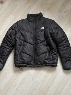 Svart pufferjacka från The North Face - Säljer en svart pufferjacka från The North Face med dragkedja framtill, hög krage och justerbar dragsko i nederkant. Jackan har logga på bröstet och ryggen samt ett klassiskt quiltat mönster. Perfekt för kalla dagar och riktigt skön att bära.