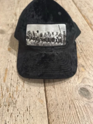 Svart truckerkeps från Hatstore - Säljer en svart truckerkeps från Hatstore med klassiskt mesh-tyg på baksidan och justerbar snapback-spänne. Kepsen har en böjd skärm och är perfekt för dig som vill ha en clean och stilren look.