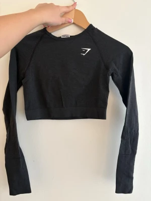 Svart crop top från Gymshark - Snygg svart långärmad crop top från Gymshark med diskret struktur i tyget och logga på bröstet. Tillverkad i stretchigt funktionsmaterial som passar perfekt till träning och gym. Tight passform och rund halsringning.