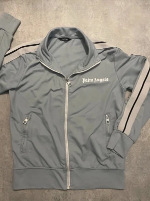 ziptröja - Ljusblå ziptröja med vita och svarta ränder längs ärmarna. Tröjan har vit dragkedja, två fickor med dragkedja och logga tryckt på bröstet. Sportig design med hög krage och ribbade muddar.