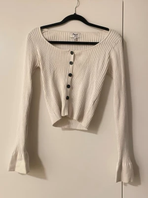 Vit ribbad kofta från fresh lions  - Snygg vit ribbad kofta från fresh lions med croppad passform och långa ärmar. Koftan har rundad urringning och svarta knappar framtill. Perfekt att styla med jeans eller kjol för en trendig look.