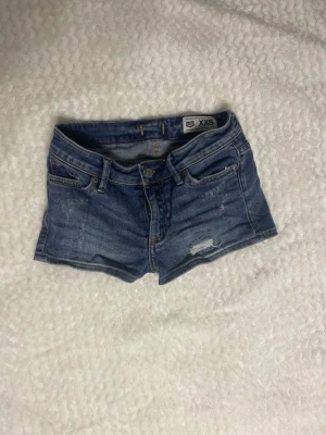 Blå denimshorts low waist - Säljer ett par blå denimshorts i klassisk femficksmodell. Shortsen har låg midja, slitna detaljer och rå kant nedtill för en avslappnad look. Perfekta för varma dagar och enkel att matcha med allt.