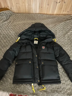 Svart dunjacka från Fjällräven - Snygg svart dunjacka från Fjällräven med gul snörning i huvan och nederkant. Fjällrävens klassiska logga på bröstet. Perfekt för kalla dagar❤️ säljer då den inte passar❤️
