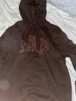 Brun hoodie från GAP med logga - Mörkbrun hoodie från GAP med stor logga i brunt och beige på bröstet. Klassisk känguruficka framtill och justerbar huva med snören. Tillverkad i mjuk bomullsblandning som känns skön mot huden. Perfekt för en avslappnad och trendig look.