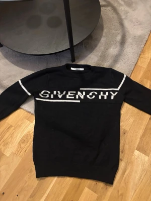 Stickad tröja givenchy small - Snygg svart stickad tröja från Givenchy med vit logga och vita linjer över bröstet och på ärmarna. Tröjan har rund halsringning och ribbade muddar vid ärmslut och nederkant. Perfekt för dig som gillar exklusiva streetwear-plagg.