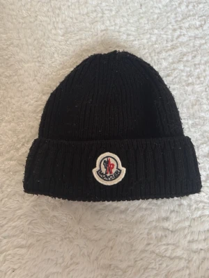 Svart ribbstickad Moncler mössa - Säljer en svart ribbstickad mössa med uppvikt kant och det klassiska Moncler-logotypen framtill. Mössan är gjord i ett mjukt och varmt material som passar perfekt för kyliga dagar. Enkel och stilren design som funkar till det mesta.
