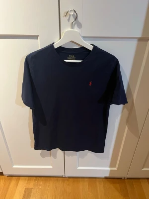 Mörkblå t-shirt från Polo Ralph Lauren - Snygg mörkblå t-shirt från Polo Ralph Lauren med klassisk passform och liten röd logga broderad på bröstet. T-shirten har rund halsringning och är tillverkad i mjuk bomull. Storlek XL i barn men passar som en M för vuxna
