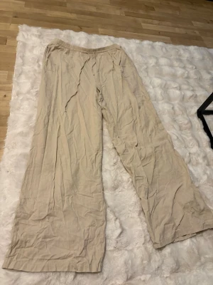 Beiga vida linnebyxor med dragsko - Snygga och luftiga linnebyxor i beige med bred passform och resår i midjan. Perfekta för en avslappnad och trendig stil. Materialet är linne som ger en skön känsla och naturlig look. Dom är lite skrynkliga på bilden, då dom legat i en låda. 