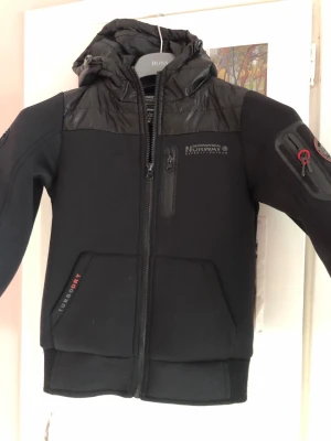 Svart vindjacka från Geographical Norway - Svart vindjacka från Geographical Norway med huva, dragkedja framtill och detaljer i syntetmaterial. Jackan har fickor med dragkedja, tryck på ärmen och bröstet samt sportig design. Perfekt för kyliga dagar och outdoor-aktiviteter.storlek för 10 år i jättebra skick