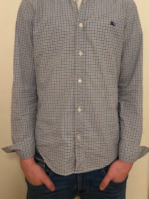Rutig Burberry skjorta  - Snygg rutig skjorta från Burberry i storlek S. Skjortan är vit, blå och brun med klassiskt rutmönster, knappar framtill och långärmad design. Tillverkad i mjuk bomull och har en broderad Burberry-logga på bröstet. Perfekt för dig som gillar stilrena och tidlösa plagg. Den är lite sliten men inget man tänker på