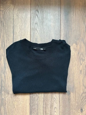 Svart stickad topp med kort ärm - En stilren svart stickad topp från 157 med rund halsringning och korta ärmar. Toppen har ribbade kanter vid hals, ärmslut och nederkant för en clean look. Perfekt att matcha med jeans eller kjol för en enkel men snygg outfit.